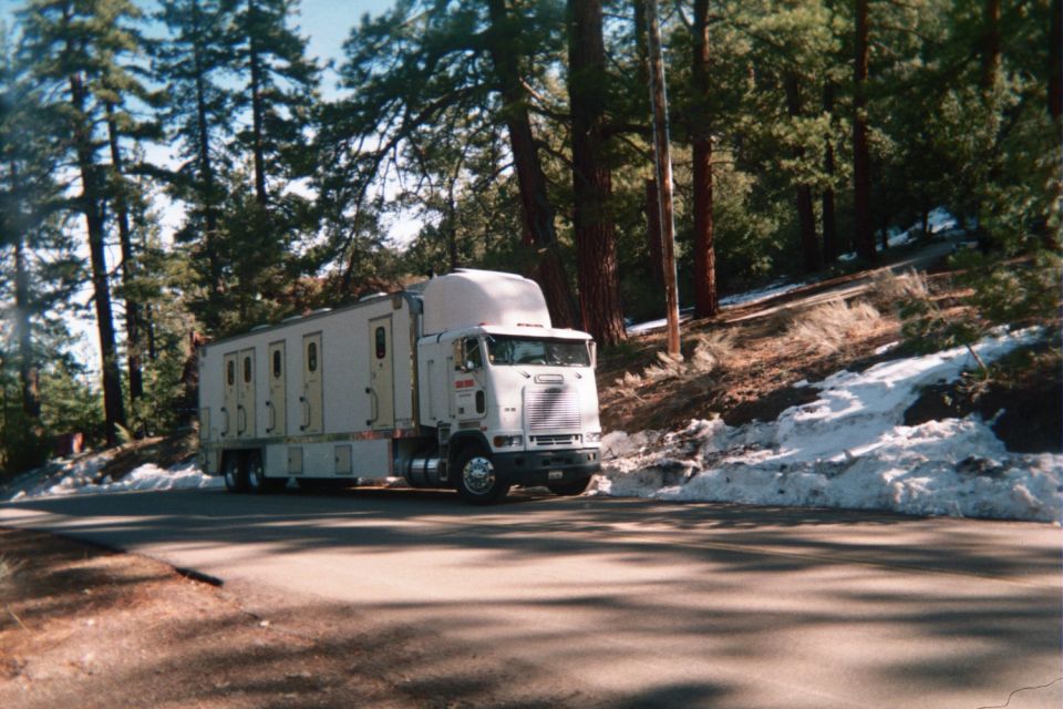 White cab-over semi with a windowed trailer parked on a forest road with patches of snow