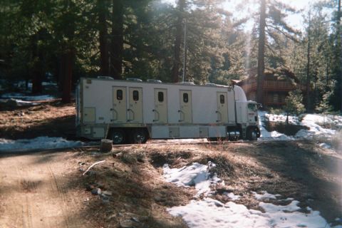 1990 Ford Bronco - long white semi trailer fitted with several side doors and windows, parked on dirt among pines with patchy snow and a cabin nearby.