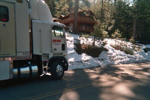 1990 Ford Bronco - white cab-over semi truck with red lettering parked on a road beside a snowy bank, with a wooden cabin among the pines.
