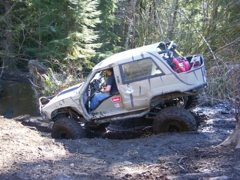 1990 Ford Bronco - This heavily modified off-road vehicle is navigating a challenging muddy terrain in a wooded area.