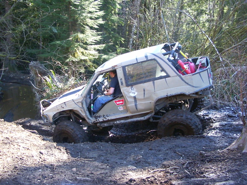 Tan off-road SUV with large tires stuck in mud on a wooded trail. Driver is visible inside