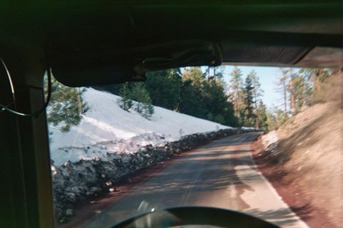 1990 Ford Bronco - Looking out a vehicle windshield at a curving rural road skirting a snow-covered slope lined with pine trees.