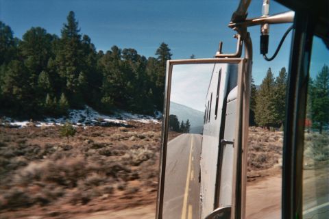 1990 Ford Bronco - vehicle side mirror reflects a trailer and the road behind while passing high-desert scrubland dotted with pine trees.