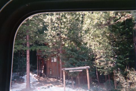 1990 Ford Bronco - Seen through a vehicle's rear window, a small wooden cabin sits among dense forest trees with patches of snow on the ground.