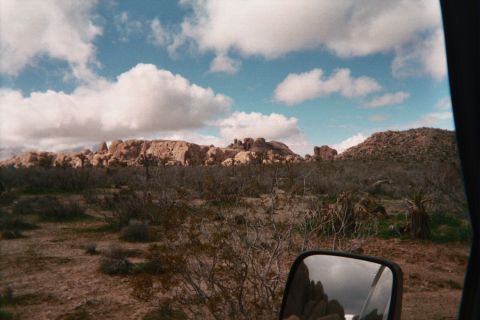 1990 Ford Bronco - high-desert landscape of scrub and rounded rock outcrops under puffy clouds, framed by a vehicle's side mirror in the corner.