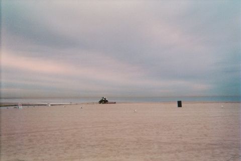 1990 Ford Bronco - vast empty beach at dusk under a pink and gray sky, with a small tractor grooming the sand near the distant waterline.