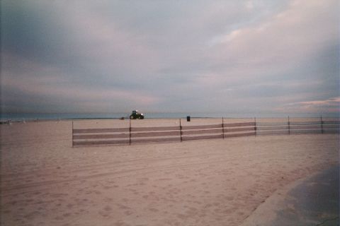 1990 Ford Bronco - empty beach at dusk lined with low slatted sand fencing, a small tractor working in the distance under heavy clouds.