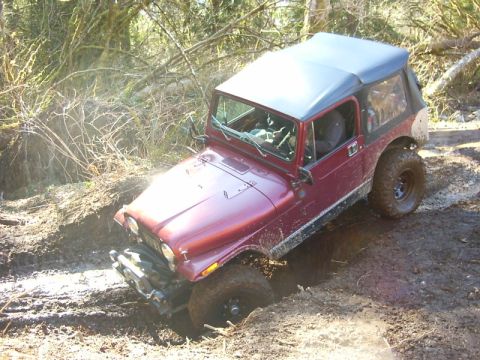 1990 Ford Bronco - A maroon Jeep CJ-7 navigates a muddy trail, its front-left wheel deeply embedded in a rut.