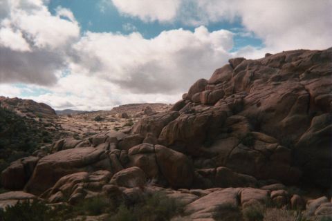 1990 Ford Bronco - massive jumble of tan boulders slopes across the desert with distant hills visible under bright puffy clouds.