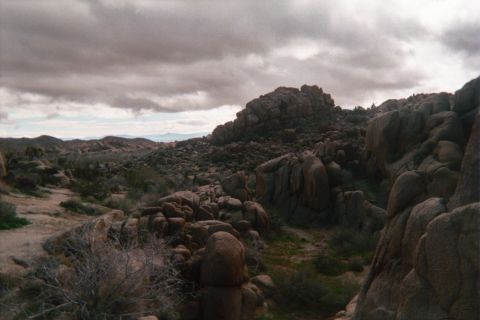 1990 Ford Bronco - boulder-covered desert ridge rises against a dark overcast sky, with low scrub and distant hills under gray clouds.
