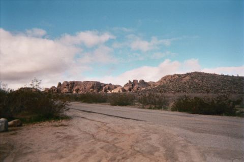 1990 Ford Bronco - paved road runs past desert scrub toward a low rocky ridge and a brushy hill beneath a bright blue sky with scattered clouds.