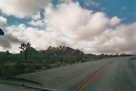 1990 Ford Bronco - desert highway with a yellow center line curves past a rock formation and Joshua trees, shot from a moving vehicle's hood.