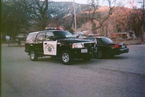 1990 Ford Bronco - black-and-white police SUV with a Highway Patrol star door shield sits beside a matching sedan on a road below wooded hills.