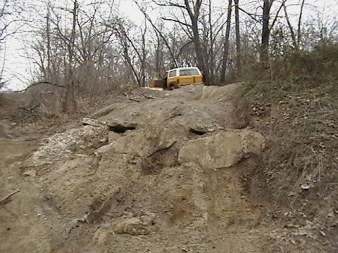 1986 Ford Bronco - A yellow and white pickup truck navigates a rocky, dirt incline in a wooded area.