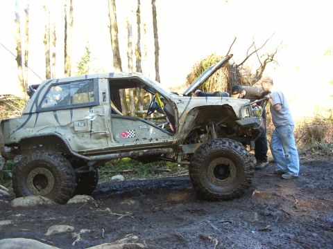 1990 Ford Bronco - Two people work on a heavily modified off-road SUV stuck in the mud on a trail.