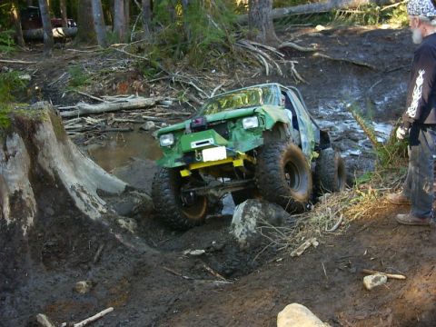 1990 Ford Bronco - A modified off-road vehicle tackles a challenging muddy terrain with a person watching.