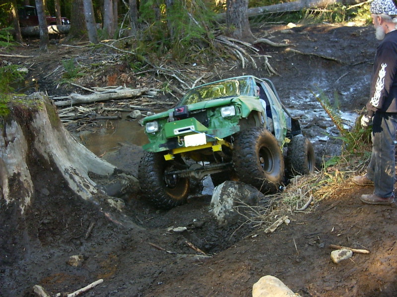 Green off-road vehicle with large tires navigating a muddy, wooded trail