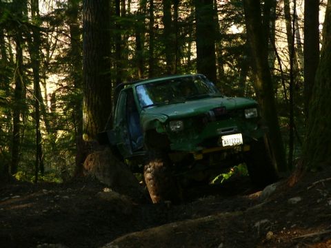 1990 Ford Bronco - A green off-road vehicle navigates a challenging wooded trail, its large tires gripping the uneven terrain.