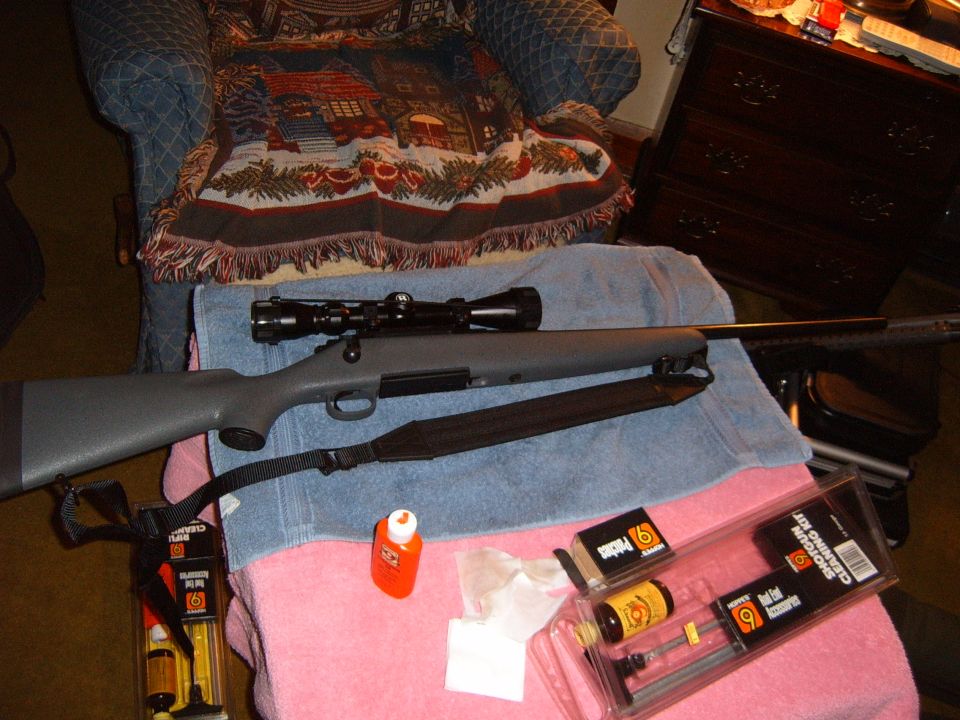 Rifle with scope and sling next to a shotgun cleaning kit and oil bottle on towels