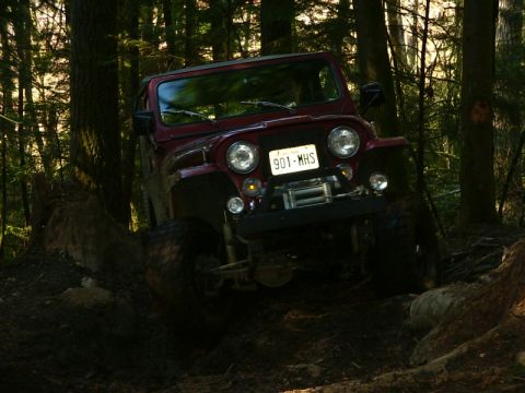 1990 Ford Bronco - A red Jeep CJ-7 navigates a challenging, muddy off-road course surrounded by dense forest.