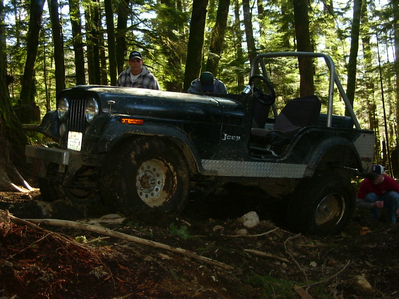 Black Jeep CJ-7 with roll cage and diamond plate trim on muddy forest trail. People visible inside and near the vehicle