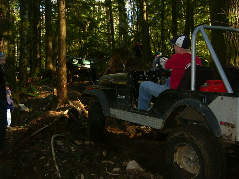 Black Jeep on a wooded trail, driver wearing a red shirt and hat. Another green Jeep is visible behind it