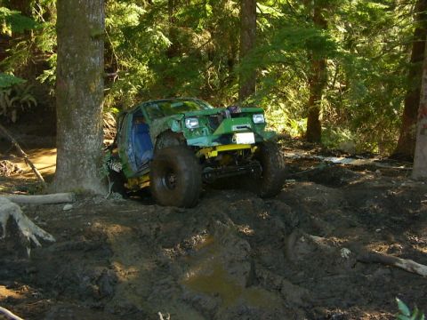 1990 Ford Bronco - A heavily modified green off-road vehicle is stuck in a muddy forest clearing.