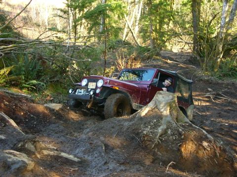 1990 Ford Bronco - A red Jeep CJ-7 tackles a challenging muddy off-road course, with its driver looking on.