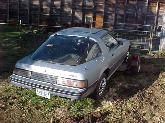 Silver-blue Mazda RX-7 coupe, rear three-quarter view, parked on grass by a barn and metal gate
