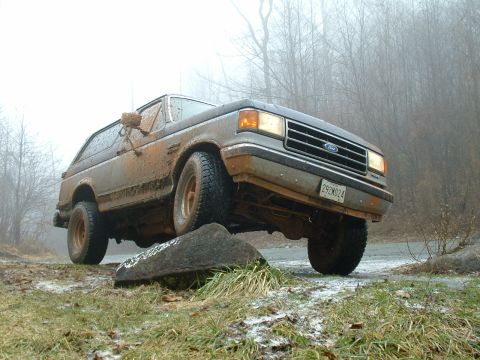 1991 Ford Bronco - A muddy 1987 Ford Bronco navigates an off-road obstacle on a foggy day.