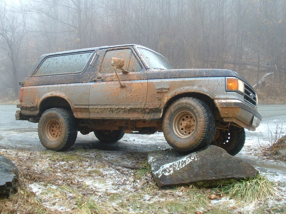Mud-caked Ford Bronco flexing with front tire on a rock, side view
