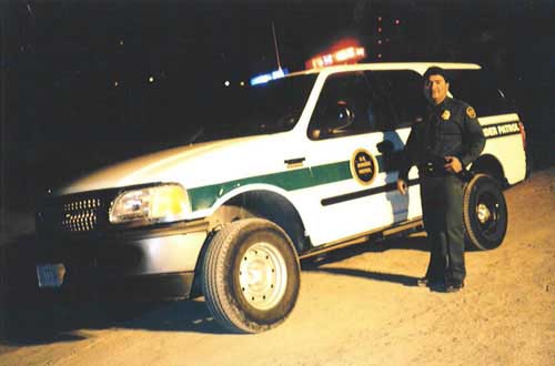White Ford Border Patrol SUV with light bar at night, uniformed agent standing alongside