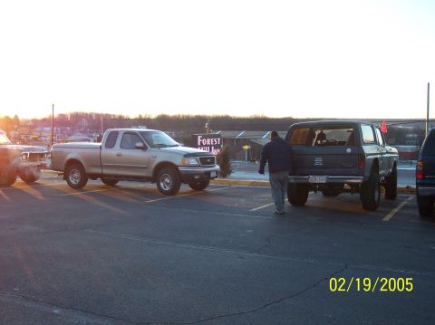 1995 Ford Bronco - A man walks away from a lifted Ford Bronco in a parking lot at sunset, with a pickup truck nearby.