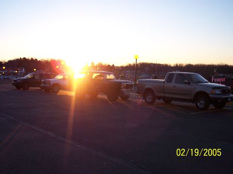 1995 Ford Bronco - Several vehicles are parked in a lot at sunset, with the sun flaring brightly.