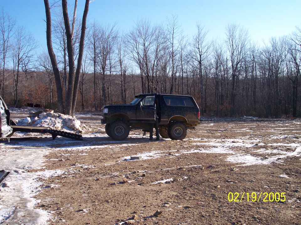Black Ford Bronco parked in a muddy, wooded area with snow patches. A trailer is attached to another vehicle on the left
