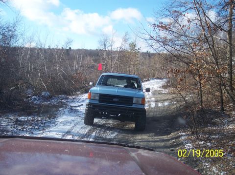 1995 Ford Bronco - Light blue pickup truck navigates a snowy dirt road in a wooded area.