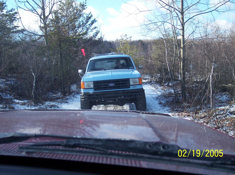 Blue Ford truck with lifted suspension driving on a snowy path, seen from another vehicle's dashboard