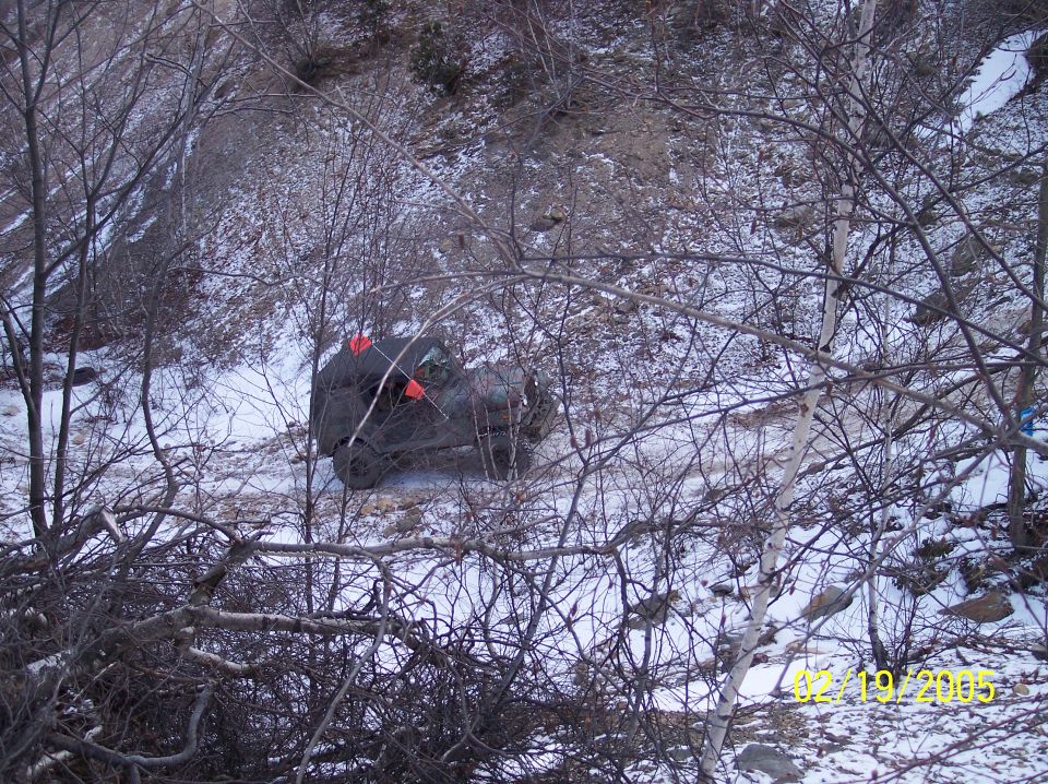 Dark green 1992 Ford Bronco overturned in snowy terrain, partially obscured by bare branches
