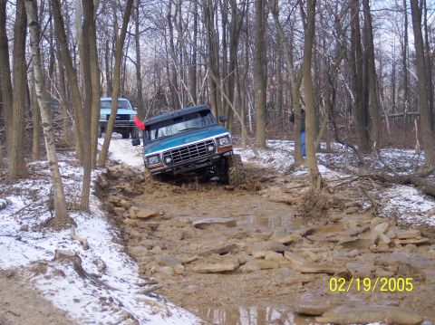 1995 Ford Bronco - Two SUVs tackle a challenging muddy and rocky path through a wooded area during winter.