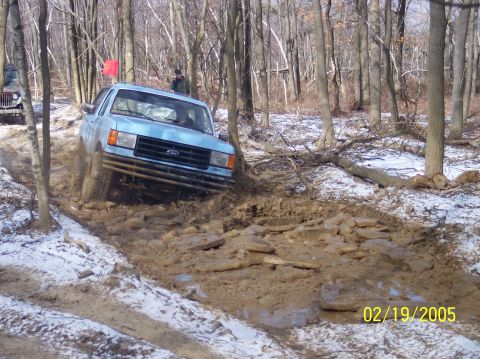 1995 Ford Bronco - A light blue 1995 Ford Bronco tackles a challenging muddy off-road course in a wooded area.