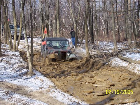 1995 Ford Bronco - A Jeep CJ-5 struggles through a deep mud pit on a wooded trail, with another vehicle visible in the background.