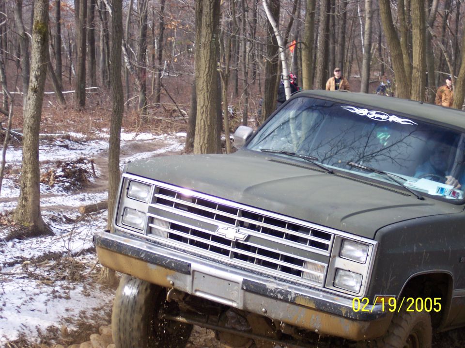 Olive green Chevrolet truck with chrome grille, off-roading in a wooded area