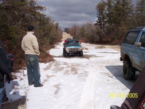 1995 Ford Bronco - Two off-road vehicles navigate a snowy path through a wooded area on a cloudy day.