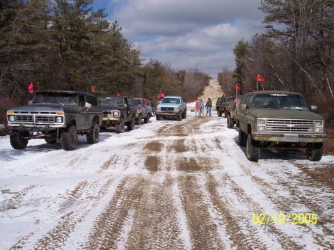 1995 Ford Bronco - A convoy of modified trucks and SUVs navigate a snowy, wooded trail on a cloudy day.