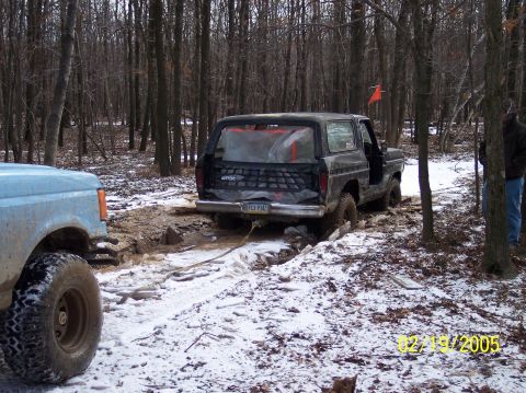 1995 Ford Bronco - Two off-road vehicles are stuck in the mud and snow in a wooded area, with one vehicle being towed.