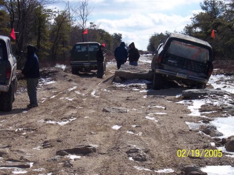 1995 Ford Bronco - Multiple off-road vehicles tackle a challenging, rocky trail in a wooded area.