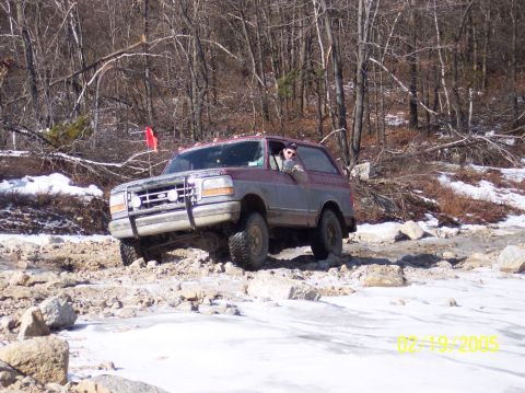 1995 Ford Bronco - A red and grey Ford Bronco navigates a rocky, snow-covered trail in a wooded area.