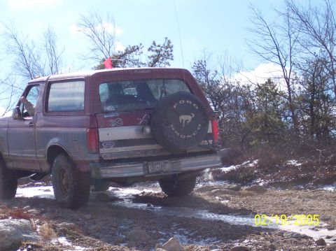 1995 Ford Bronco - A 1995 Ford Bronco navigates a muddy and rocky path, with a spare tire mounted on the back.