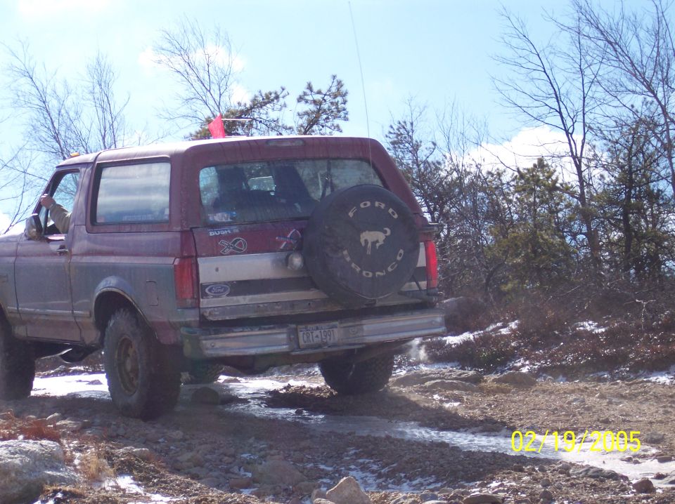 Rear view of a 1995 Ford Bronco driving on a rocky, wet trail with trees in the background