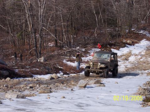 1995 Ford Bronco - A Jeep CJ-7 navigates a challenging icy and rocky path in a wooded area, with a person observing the off-road excursion.